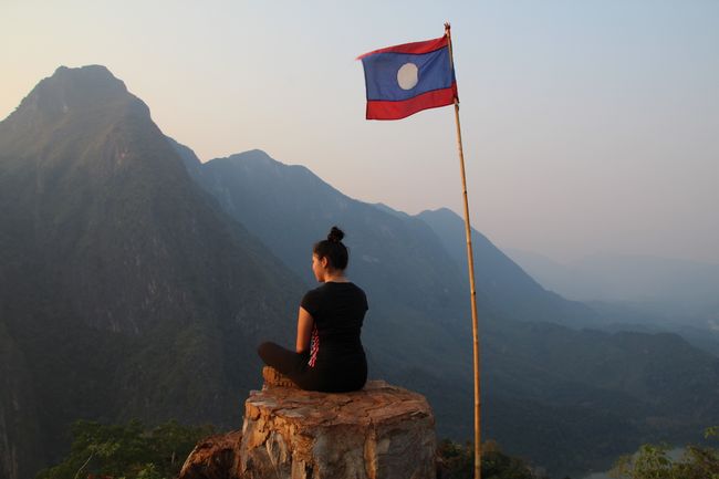 Franzi sitzt neben der laotischen Flagge vor gigantischen Bergen