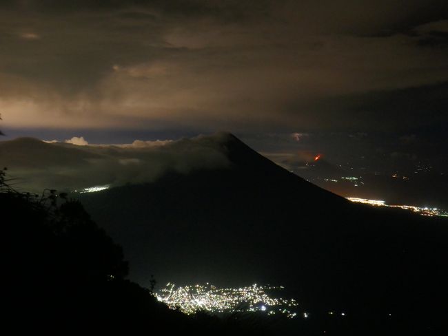 Guatemala City illuminating the sky, Antigua on the left under the clouds at the foot of Agua. In the background on the right, a thunderstorm and the glowing lava flow of Pacaya volcano